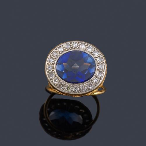  Anillo con centro de zafiro oval sintético y orla de diamantes talla 8/8 en montura de oro amarillo de 18K y vista en platino.