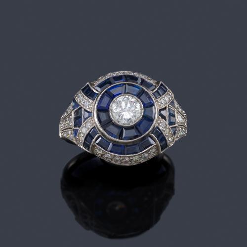Lote 2072
Anillo estilo 'art decó' con brillante central de aprox. 0,60 ct con orla y bandas de zafiros calibrados, enriquecidos con diamantes de aprox. 0,70 ct en total.