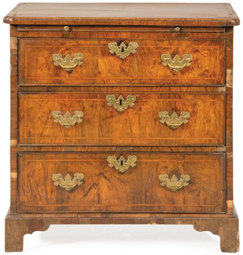 Lote 1102
Cómoda Jorge I en madera de nogal, con tapa moldurada con balda deslizante a modo de escritorio, sobre tres cajones con tiradores y escudos de cerradura en latón recortado. 
Inglaterra, segundo tercio S. XVIII