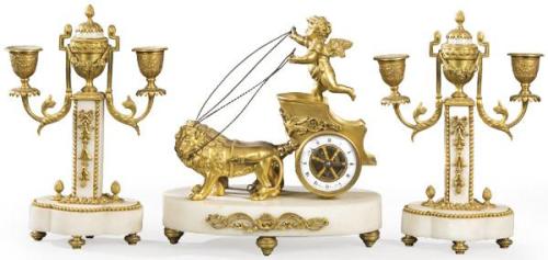  Reloj de sobremesa Luis XVI con guarnición de pareja de candelabros. Sobre basamento  ovalado de mármol blanco con aplicaciones de bronce sobre el que se sitúa una biga romana tirada por dos leones.