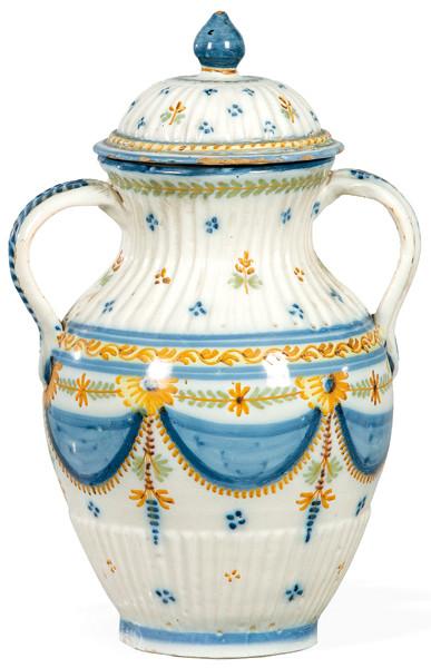 Jarrón con tapa, cuerpo ovoide, cuello y base gallonados. Sobre esmalte de fondo blanco, con dibujo perfilado en negro y colores azul celeste, verde oliva, ocre y amarillo; representando guirnaldas y cortinajes.