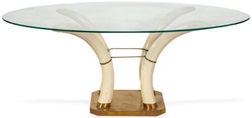 Mesa de comedor con tapa de vidrio ovalada, sobre cuatro colmillos de resina imitando marfil, apoyados en una plataforma de latón.