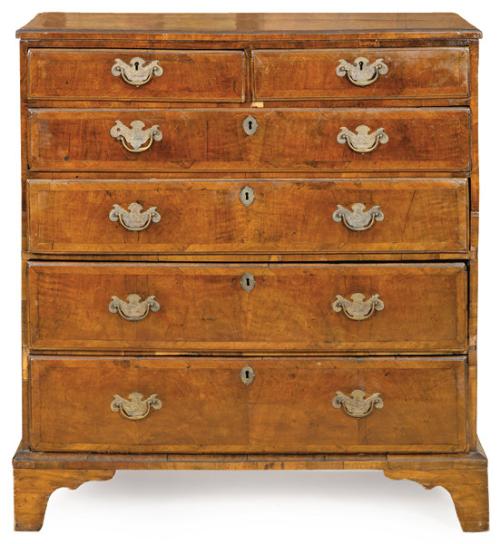 Cómoda Jorge I en madera de nogal, con tapa moldurada sobre seis cajones con tiradores y escudos de cerradura en latón recortado y grabado. Inglaterra, h. 1720