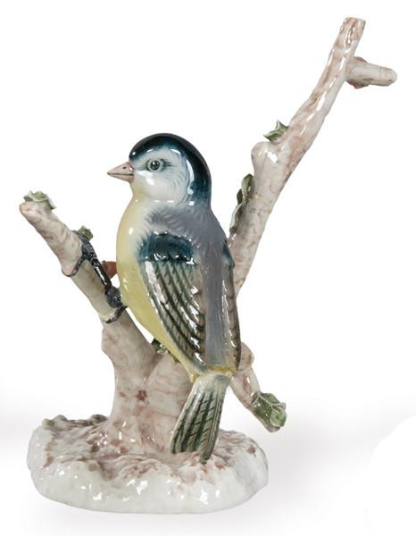  Figura de ave en porcelana policromada, manufactura centroeuropea. Composición con ave posada sobre tronco estilizado y base orgánica. Decoración bajo cubierta en tonos amarillos, azules y grises.