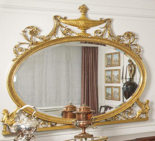 Espejo ovalado eduardino siguiendo modelos Adam con decoración tallada y dorada, rematado por una urna en forma de copa.
