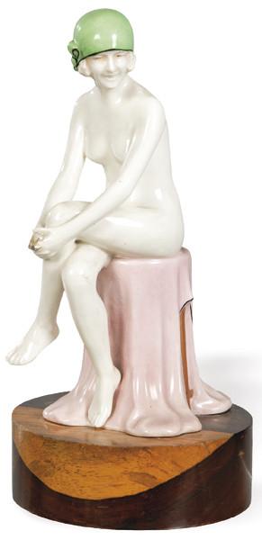 Figura de bañista Art Decó en cerámica esmaltada probablemente Royal Dux , h. 1925-1935, representada sentada sobre paño rosa, con gorro verde y cuerpo en esmalte blanco marfil. Sobre base circular de madera posterior.