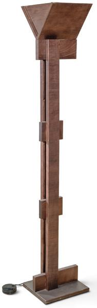 Lámpara de pie con estructura arquitectónica en madera teñida, con base prismática, fuste vertical macizo, dos resaltes laterales de unión y remate superior facetado en forma de copa invertida. 